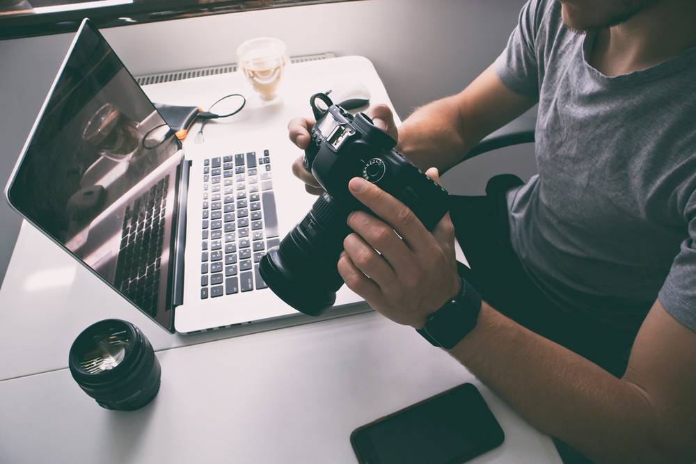 Photographer holding camera at desk with laptop | Skylum Blog