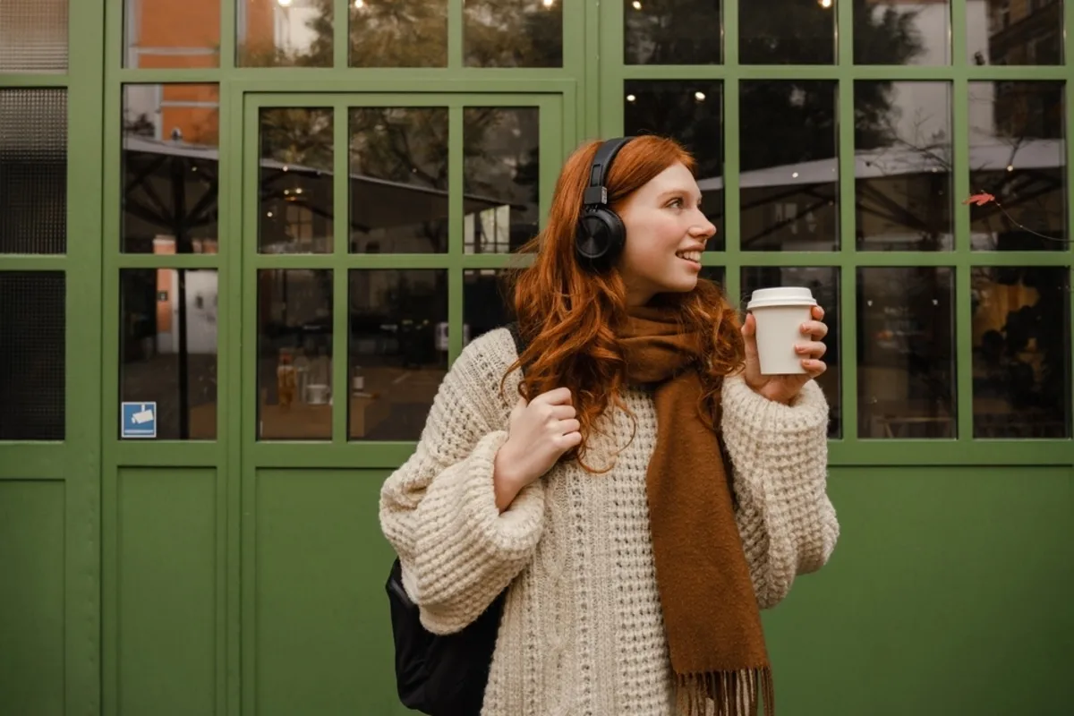 A girl in a scarf and headphones drinking coffee near a building | Skylum Blog