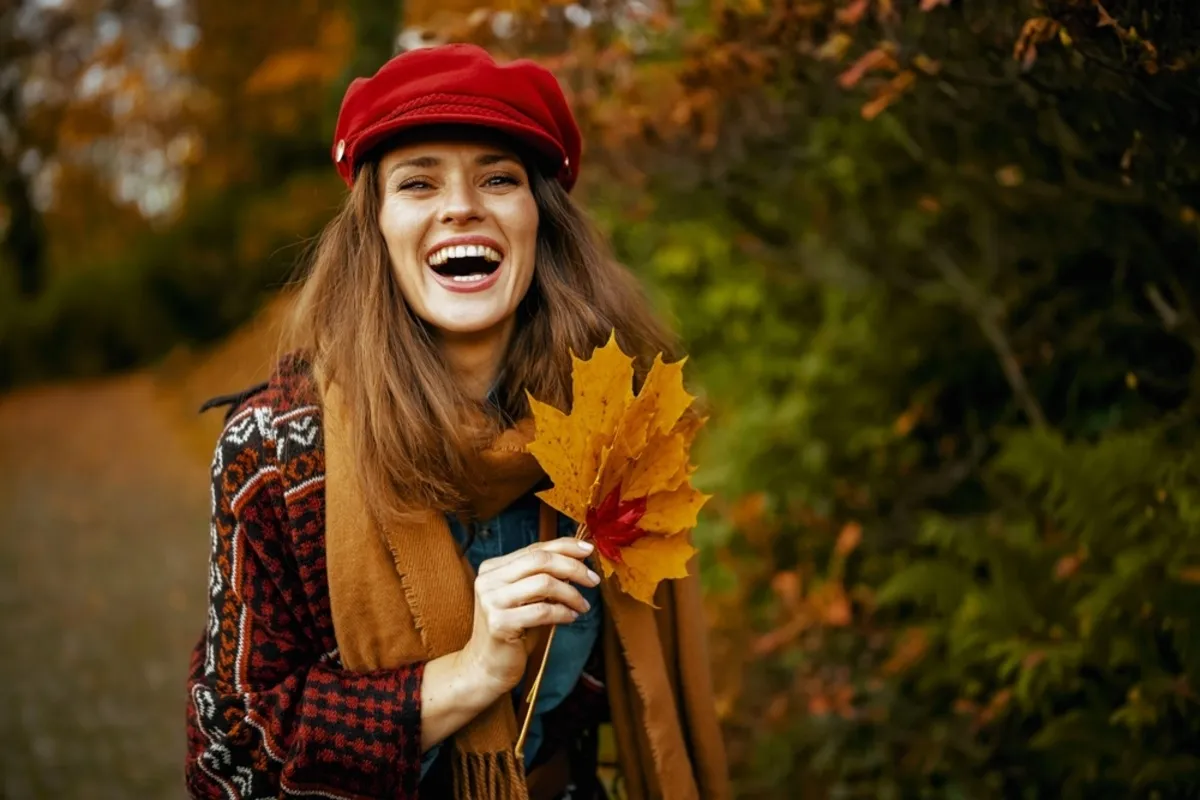 A girl in a hat and scarf holds autumn leaves and smiles | Skylum Blog