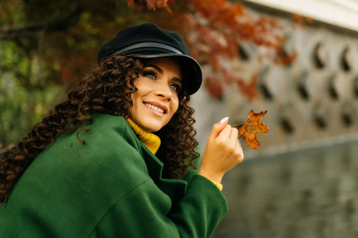 A girl in a green coat and hat holds an autumn leaf in her hands | Skylum Blog