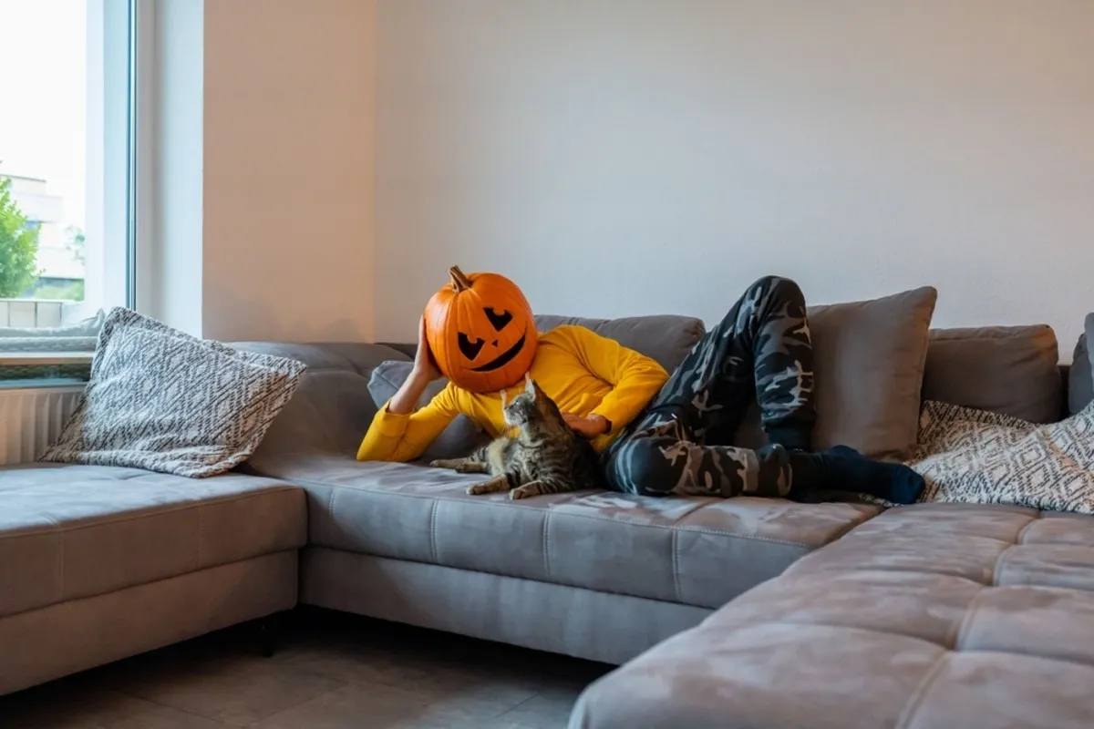 A man with a pumpkin on his head lies on the couch with a cat | Skylum Blog