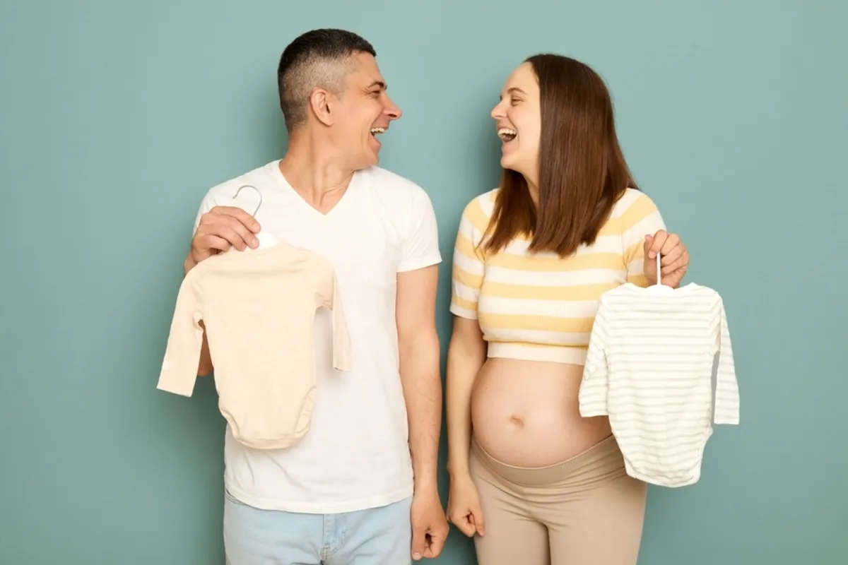 The expectant parents hold up the baby's clothes and laugh | Skylum Blog
