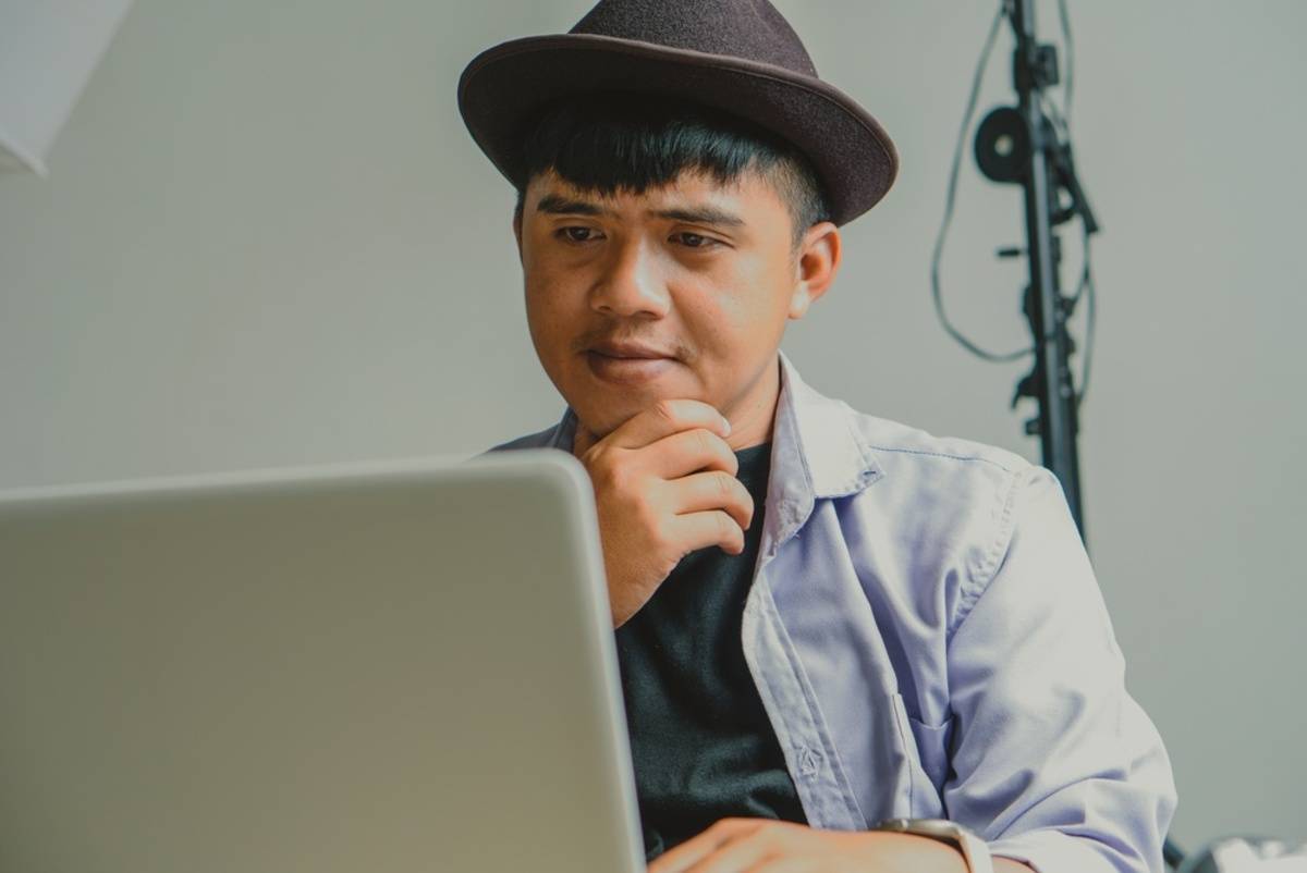 Man in a hat resting chin on hand while studying a laptop screen in a studio