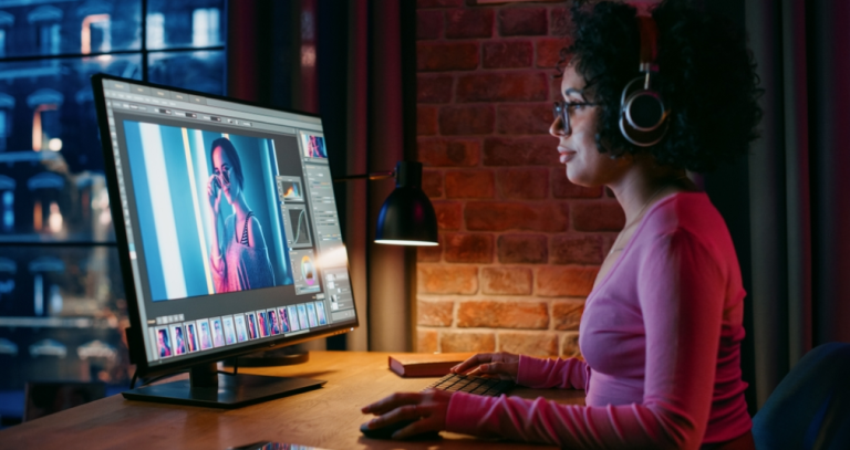Woman with headphones editing a blue-toned portrait on a desktop monitor at a brick-wall desk