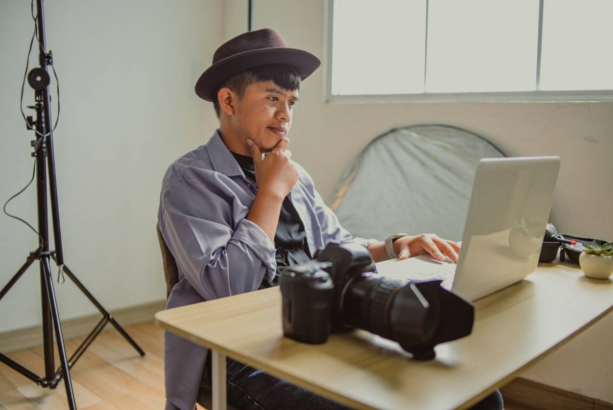 Photographer in a hat resting his chin on his hand while reviewing photos on a laptop beside a DSLR camera