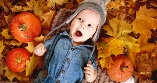 Baby in denim overalls and knit hat lying among fall leaves with small pumpkins