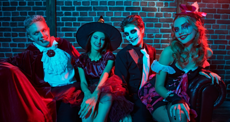 Four friends in vampire, witch and skeleton makeup costumes sit on a couch under red and blue light