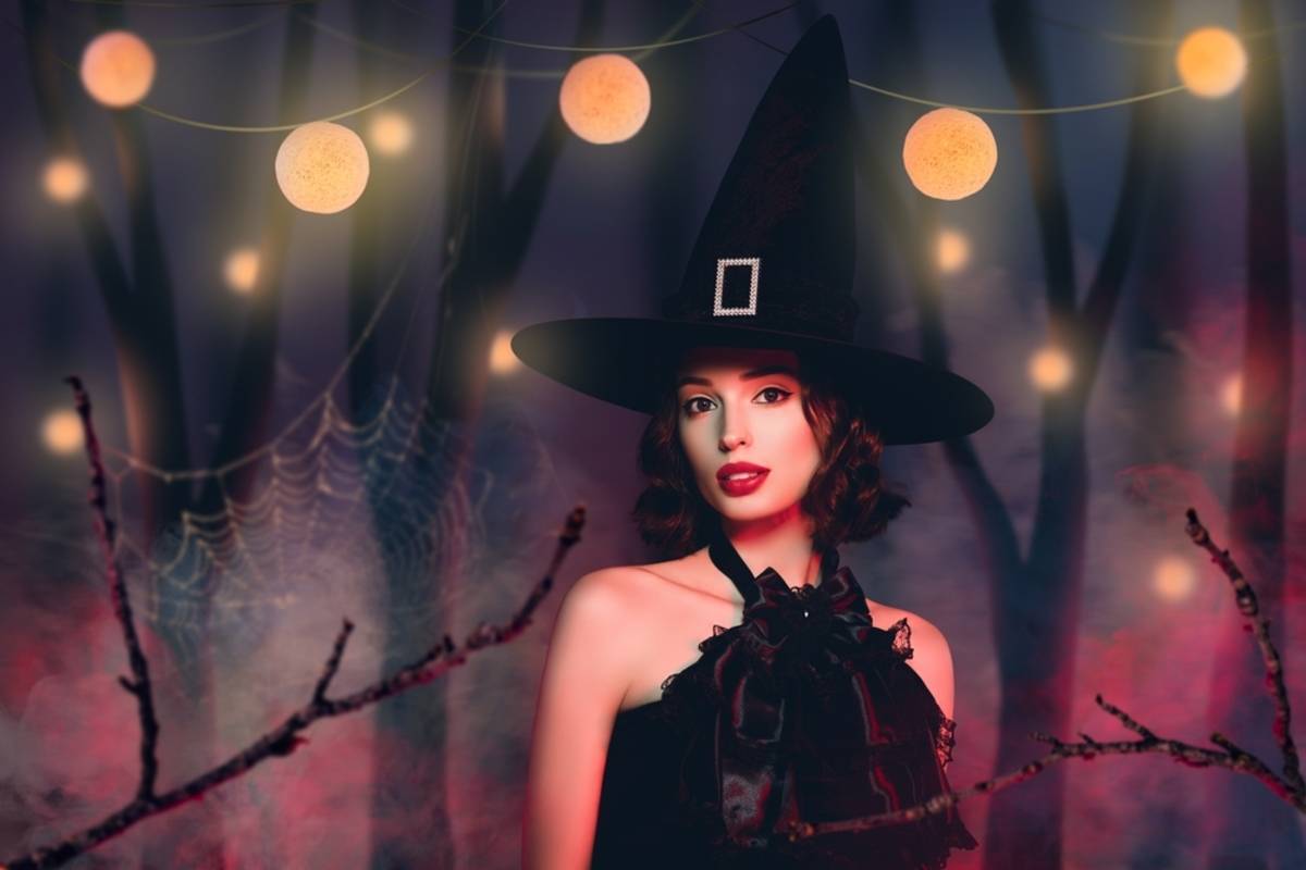 Woman in a witch hat and black ruffled collar poses in a misty forest strung with glowing orb lights