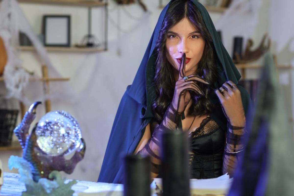 Woman in a dark green hooded cloak poses as a fortune teller beside a crystal ball on claw feet