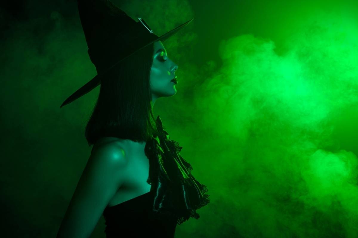 Woman in a pointed witch hat profiled against green smoke lit from one side