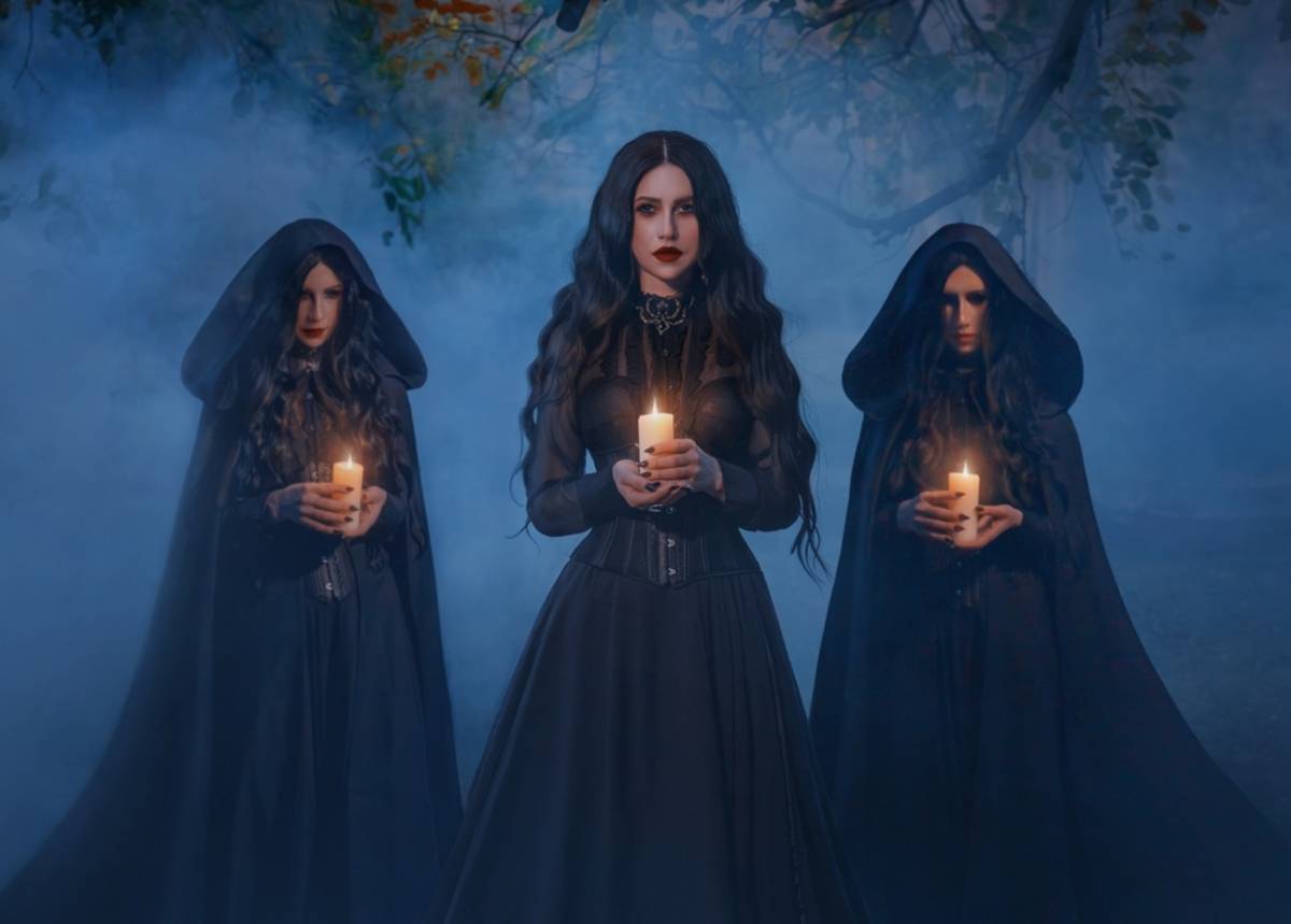 Three women in dark hooded cloaks hold lit candles in a misty forest at dusk