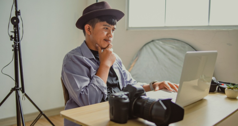 Photographer in a hat resting chin on hand while reviewing images on a laptop, camera on desk