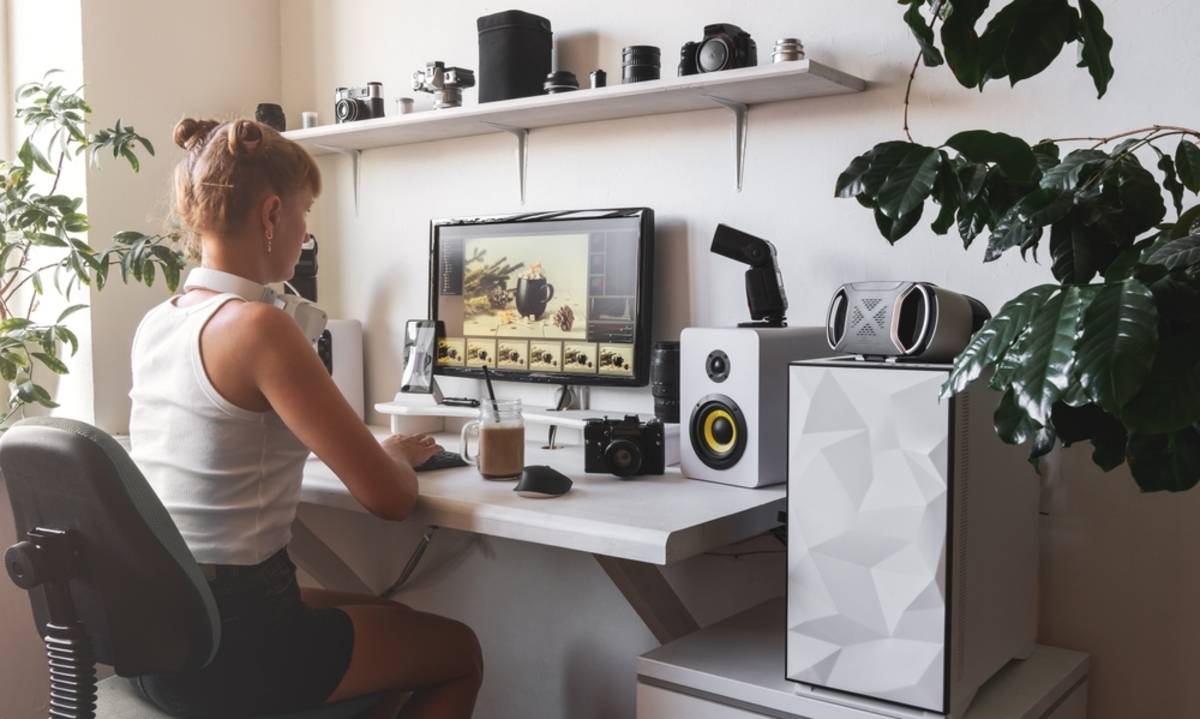 Editor at a desk reviewing a photo thumbnail strip on a monitor, surrounded by cameras and lenses