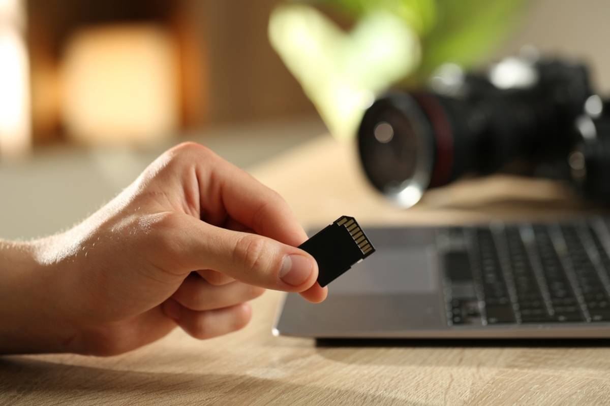 Hand holding an SD memory card near a laptop keyboard with a camera lens in the background