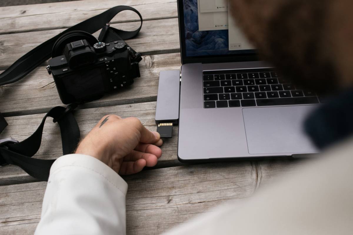 Hand inserting an SD card into a laptop reader beside a mirrorless camera on a wooden table