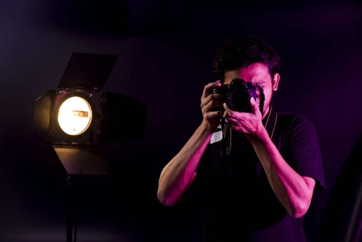 Photographer raising a camera to his eye lit by a barn-door studio lamp in a dark room