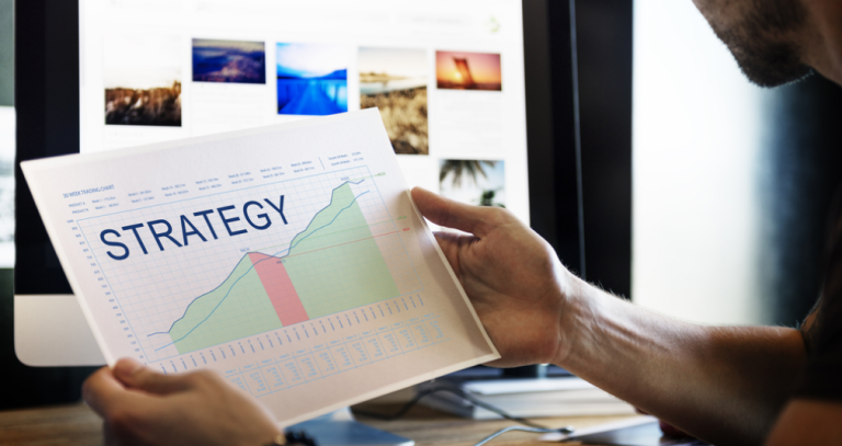 Hands holding a printed strategy chart in front of a monitor showing a photo gallery grid