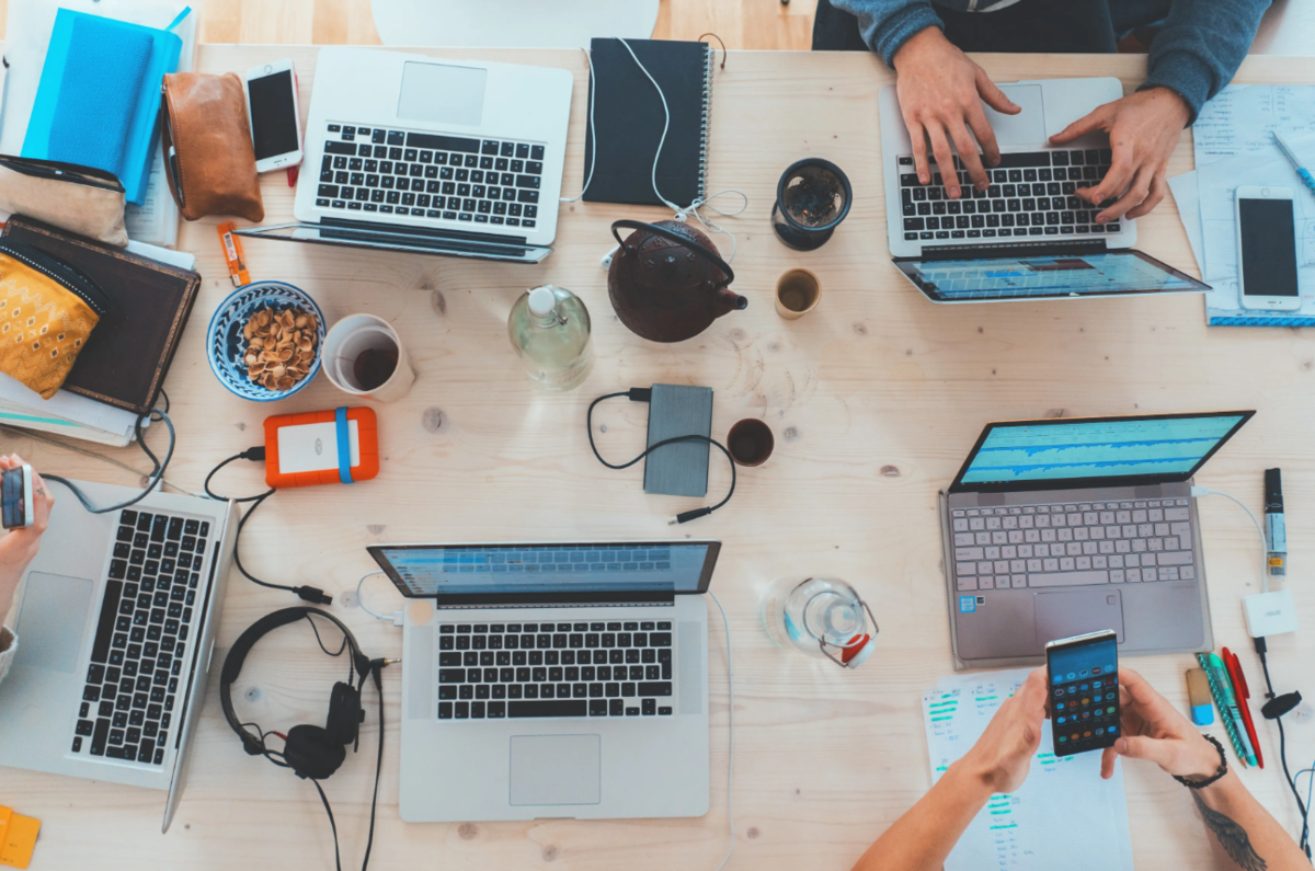 Overhead view of a desk with four laptops, phones, hard drives, headphones and coffee cups