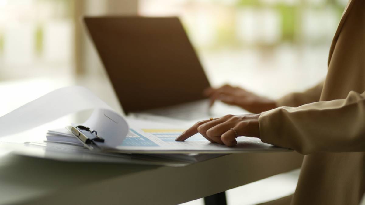 Hand on a printed chart beside a laptop keyboard, with a clipboard of papers on the desk