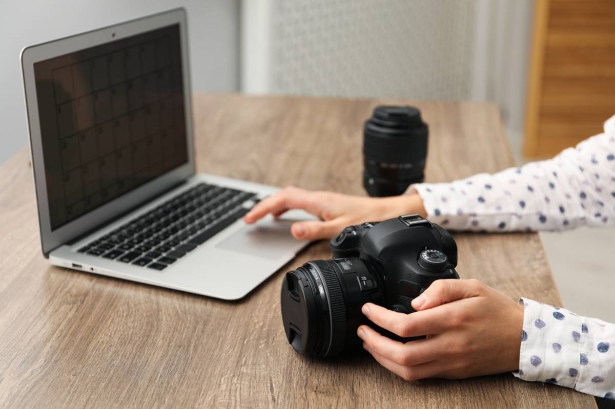 Hands holding a DSLR camera and resting on a laptop trackpad with a lens on the desk